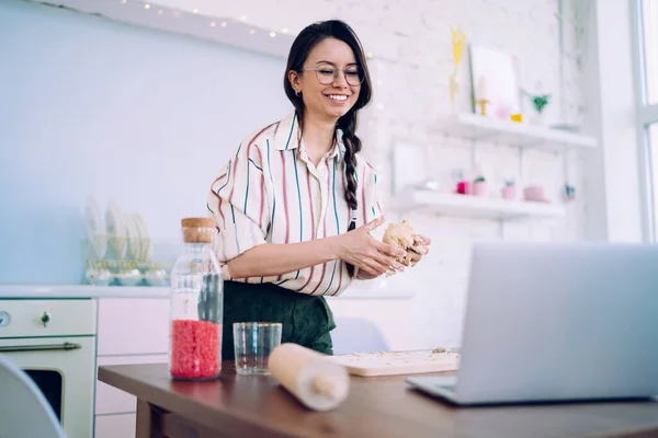 Young housewife with beautiful smile standing at table holding dough in hands while watching culinary class at laptop in comfortable kitchen