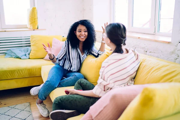 African American woman with delightful smile gesturing while telling exciting story to female friend relaxing on yellow cozy sofa at home
