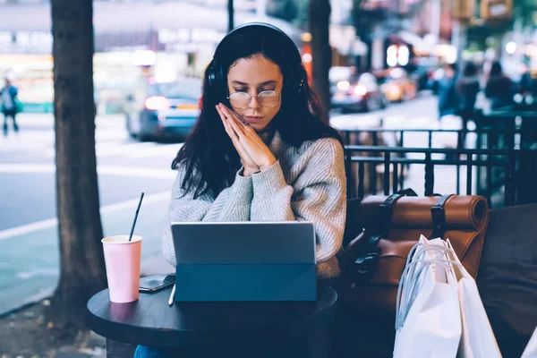 Young beautiful thoughtful slender female freelancer in glasses and in headphones with folded hands near face using tablet at table outside of cafe