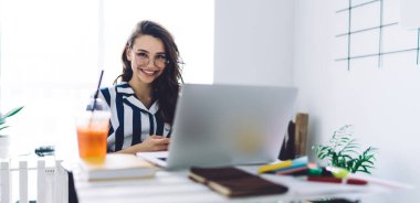 Çizgili, uzun gömlekli, yuvarlak gözlüklü, gülümseyen ve laptopuyla beyaz bir ofiste masada oturan koyu renk saçlı genç bir kadın.