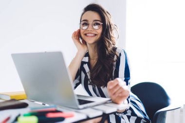 Gözlüklü ve zarif çizgili elbiseli çekici genç bir kadın ev ofisinin masasında oturmuş elinde dizüstü bilgisayarla sevinçle uzaklara bakıyor. 