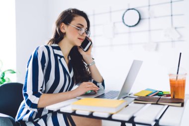 Koyu renk saçlı, gözlüklü, sarı defter ve smoothie ile masada oturan ve cep telefonuyla konuşurken bilgisayarla çalışan kadın.