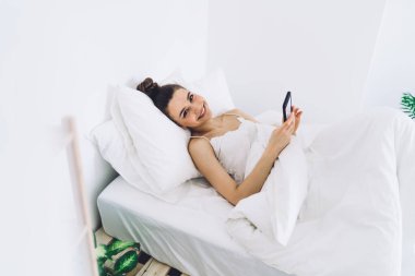Yukarıdaki mutlu kadın beyaz yastık ve battaniyeyle yatakta dinleniyor ve akıllı telefon kullanıyor ve kameraya bakıyor.