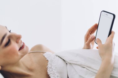 Pijamalı, gülümseyen bir kadın beyaz duvarlı minimalist yatak odasında yatarken telefonuna beyaz ekranla mesaj atıyor.