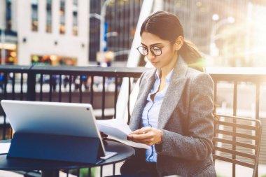 Kürklü ve gözlüklü başarılı bir etnik iş kadını sokak terasında oturmuş, New York 'ta yanan tablette sörf yaparken dokümanları okuyor. 