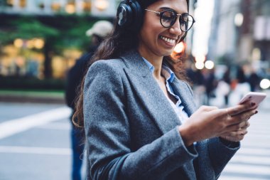 Gülümseyen esmer, etnik bir kadın. Kulaklıklı, gözlüklü, gri montlu. New York caddesinde akıllı telefonları dinliyor. 