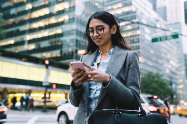 Gözlüklü, siyah çantalı, gri montlu, esmer bir kadın. New York caddesinde, bulanık arka planda akıllı telefonları dinliyor.