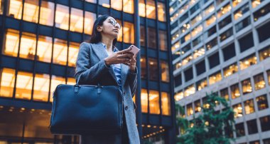 New York 'taki modern ofis binasının önünde siyah çantalı ve cep telefonlu, gri ceketli, etnik kökenli iş kadını bekliyor.