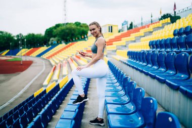 Saçları örgülü gülümseyen sarışın bir kadın ve modern spor kıyafetleri içinde boş stadyum tribünlerinde kameraya bakarken akıllı telefondan mesajlaşırken.