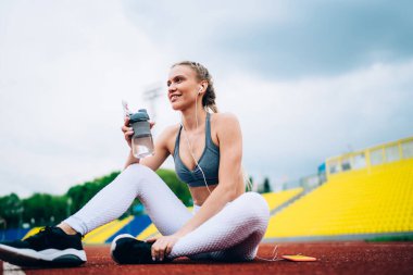Akıllı telefondan müzik dinleyen ve elinde su şişesi tutan gülümseyen bayan sporcunun zemin kattaki manzarası boş stadyumun raylarında otururken gözlerini kaçırıyor.