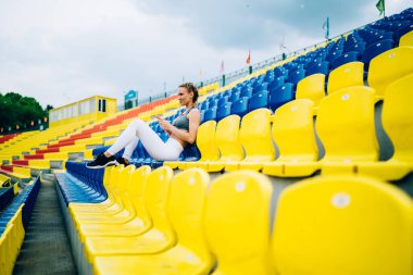 Beyaz taytlı, beyaz saçlı, gri bluzlu ve akıllı telefon kullanırken gözlerini kaçıran zayıf bayan sporcunun yan görüntüsü