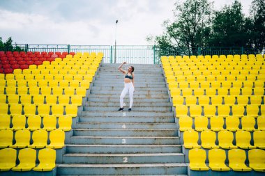 Bulutlu bir günde stadyumda kol kaslarını gösterirken sayılı basamaklarda duran rahat taytlı ve üst basamaklarda duran çekici bir kadın.