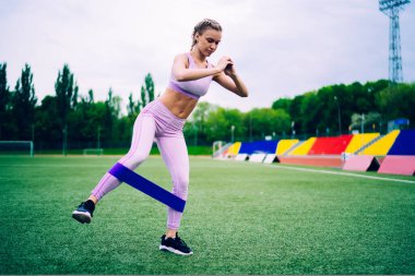 Parlak spor giyim giymiş genç bir kadın bir bacağının üzerinde dengede duruyor yeşil çimlerde direniş bandıyla antrenman yapıyor şehir stadyumunun tribününe bakıyor. 