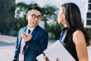 İş konferansında sokakta yürürken ve kahve molası verirken akıllı telefon kullanan modaya uyan Asyalı bir adam etnik bayan meslektaşıyla sohbet ediyor.