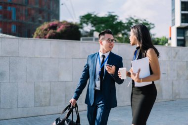 Kahveli ve gazeteli moda bir kadın güneşli bir sokakta yürüyor ve zarif Asyalı meslektaşlarıyla iş konferansında konuşuyor.