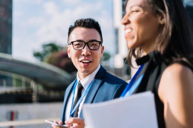 İş konferansında etnik kadın iş arkadaşıyla sokakta yürürken kameraya gülümseyen takım elbiseli yakışıklı Asyalı adam. 