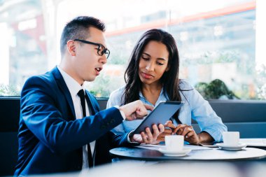 Afro-Amerikalı bir kadınla birlikte genç Asyalı bir adam kafeteryada kahve içerken tablet paylaşıyor iş görüşmesi yapıyor.