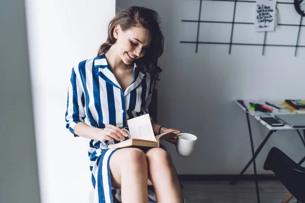 心满意足 衣冠楚楚的女士 在办公室的窗台上一边看书一边喝茶 脸上挂着灿烂的笑容 — 图库照片