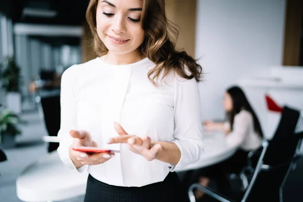 穿着正式服装的年轻貌美的女性穿着时髦的智能手机 快乐地站在工作场所的桌旁 背景模糊 — 图库照片