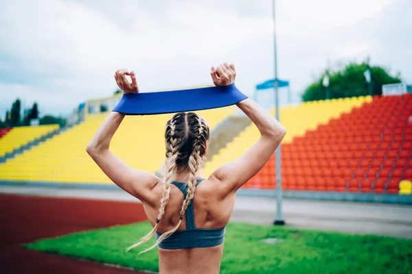 スタジアムで青いゴムリボンで熱心に訓練しながら立ち上がって腕を上げる顔のないスポーティーな女性の背景ビュー — ストック写真