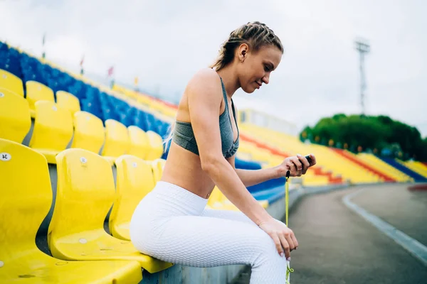 現代のスポーツ衣装で大人の運動女性のサイドビューストップウォッチで時間を見てカラフルな空のスタジアムで椅子に座って — ストック写真