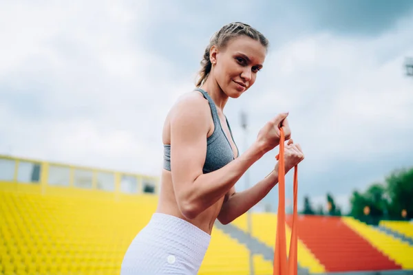穿着运动服的年轻健康而自信的女人在五颜六色的体育场做伸展运动 在模糊的背景下看着镜头 — 图库照片