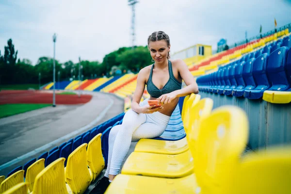 大人現代女性でスポーツウェアサーフィンスマートフォンで座っているクロス足で椅子の上にグランドスタンドでカメラを見て — ストック写真