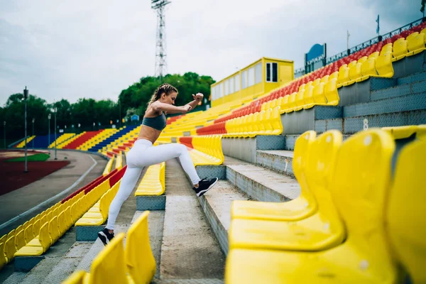 サイドビューのフィット集中女性で ピグテールは階段の上をジャンプするための演習を行います上のTriuneでスタジアムを見下ろします — ストック写真