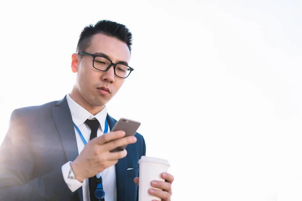 From below of modern Asian man in suit and glasses surfing phone while holding disposable cup of coffee standing in back lit on street