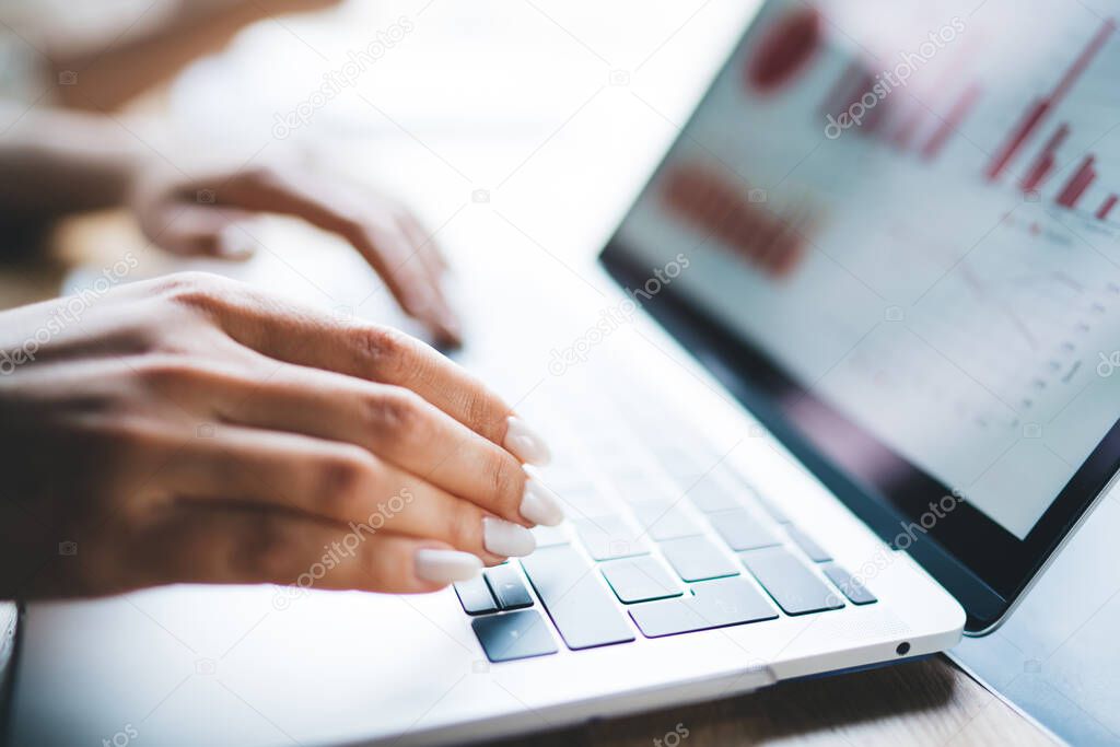 Female hands with white manicure typing on laptop keyboard with various graphs, charts and diagrams on screen and with bright light on background