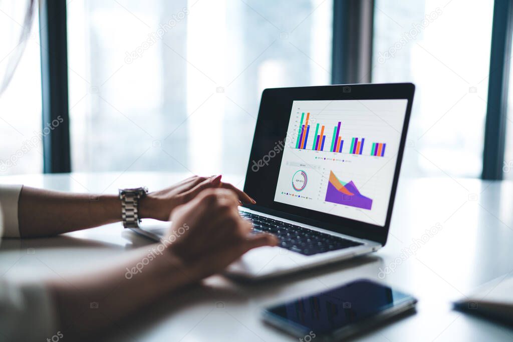 Hands of anonymous female office worker typing on keyboard of modern laptop with colorful graphs while sitting at table in office