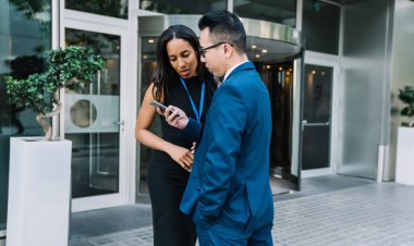 Sokakta modern binaya karşı duran ve konuşurken cep telefonunu paylaşan çok ırklı genç kadın ve erkek.