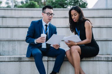 Çok ırklı bir kadın ve kahve bardaklı bir adam sokakta beton bir tribünde oturuyor ve takım halinde çalışan kağıt dökümanları tartışıyorlar.
