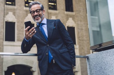 Mavi takım elbiseli ve kravatlı orta yaşlı işadamı gri ofis binasına karşı şeffaf balkonda duruyor ve cep telefonu kullanıyor.