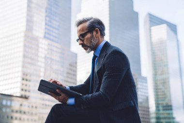 New York 'ta gökdelenlerle arka planda modern dijital tablete göz atan orta yaşlı, ciddi meşgul bir erkeğin yan görüntüsü