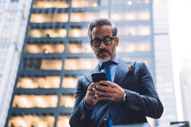 Gözlüklü ve koyu renk takım elbiseli orta yaşlı bir işadamının New York 'taki ofis binasının arka planında telefonla yazı yazması.