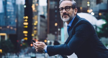 Gözlüklü ve takım elbiseli orta yaşlı bir adam elinde telefonla cam çitlere yaslanıyor ve New York caddesinin arka planına yaslanıyor.