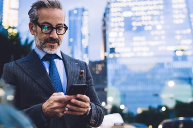 Gözlüklü ve takım elbiseli yakışıklı bir erkek telefonda yazı yazıyor ve New York karayolu ve gökdelenlerin arka planında duruyor.