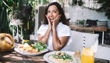 Kahvede servis edilen yemek miktarı karşısında heyecanla kameraya bakarken yanaklarından el ele tutuşan mutlu yetişkin kadın.