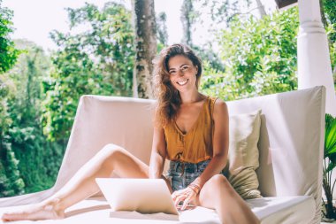 Güzel gülümseyen, neşeli, sıradan genç bir kadın yaz günü yeşil terasta rahat bir kanepede otururken dizüstü bilgisayarda geziniyor.