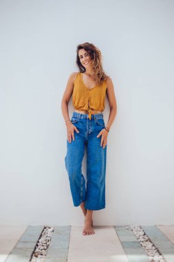 Zarif, bronzlaşmış, sarı yazlık bluzlu, ayakkabısız kadın ve beyaz arka planda duvarın kenarında duran kot pantolon ve kameraya bakıyor.