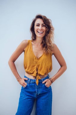 Uzun kıvırcık saçlı, kot pantolonlu, turuncu gömlekli, elleri ceplerinde gri duvarda gülümseyen çekici bir kadın. 