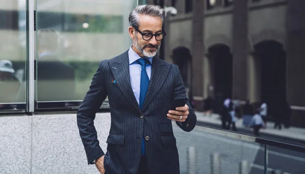 Busy serious middle aged businessman in glasses and with hand in pocket using mobile phone while standing on transparent balcony