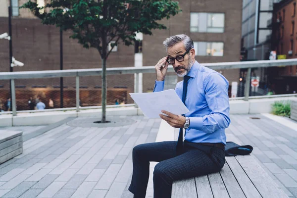 Side view of middle aged man in business suit correcting glasses while sitting on wooden bench and analyzing papers on New York street