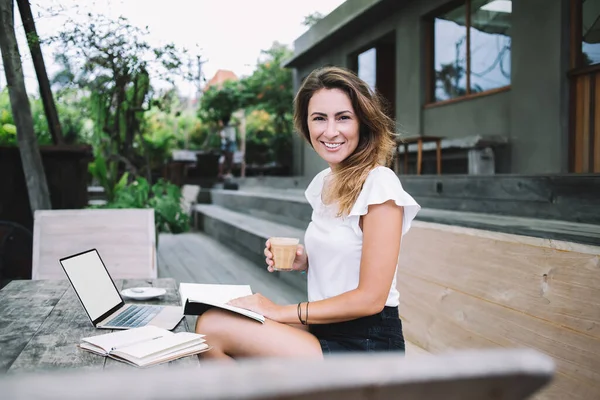 身穿休闲装 阅读书籍和喝咖啡的美丽微笑的女人 坐在木桌上 拿着笔记本电脑和笔记本 看着相机 — 图库照片
