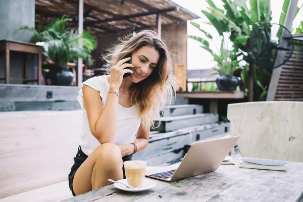 夏天的一天 身穿白色T恤和黑色短裤的年轻貌美的女人坐在桌旁 一边用手机说话 一边看笔记本电脑 — 图库照片
