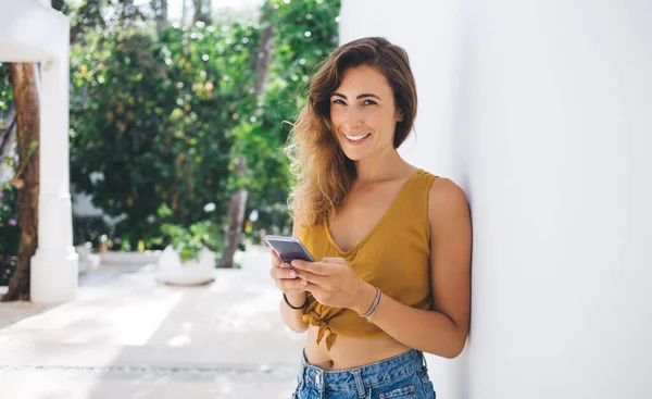 Carefree pretty woman in crop top leaning on whitewashed wall and smiling while texting on smartphone and looking at camera on blurred background with garden