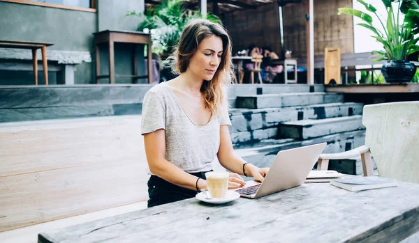 穿着休闲装的现代女性一边坐在长椅上一边吃着餐垫一边在咖啡店里畅饮 一边浏览着现代笔记本电脑 — 图库照片