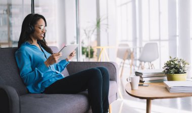 Yandan gülümseyen, gözlüklü, parlak ofisindeki koltukta oturan ve kulaklıkla tablet izleyen Afro-Amerikan çalışanın görüntüsü.