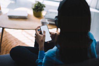 Modern bir kadının arka planda gündelik kıyafetleriyle kanepede oturup müzik dinlerken evde boş ekranla cep telefonuna bakması. 